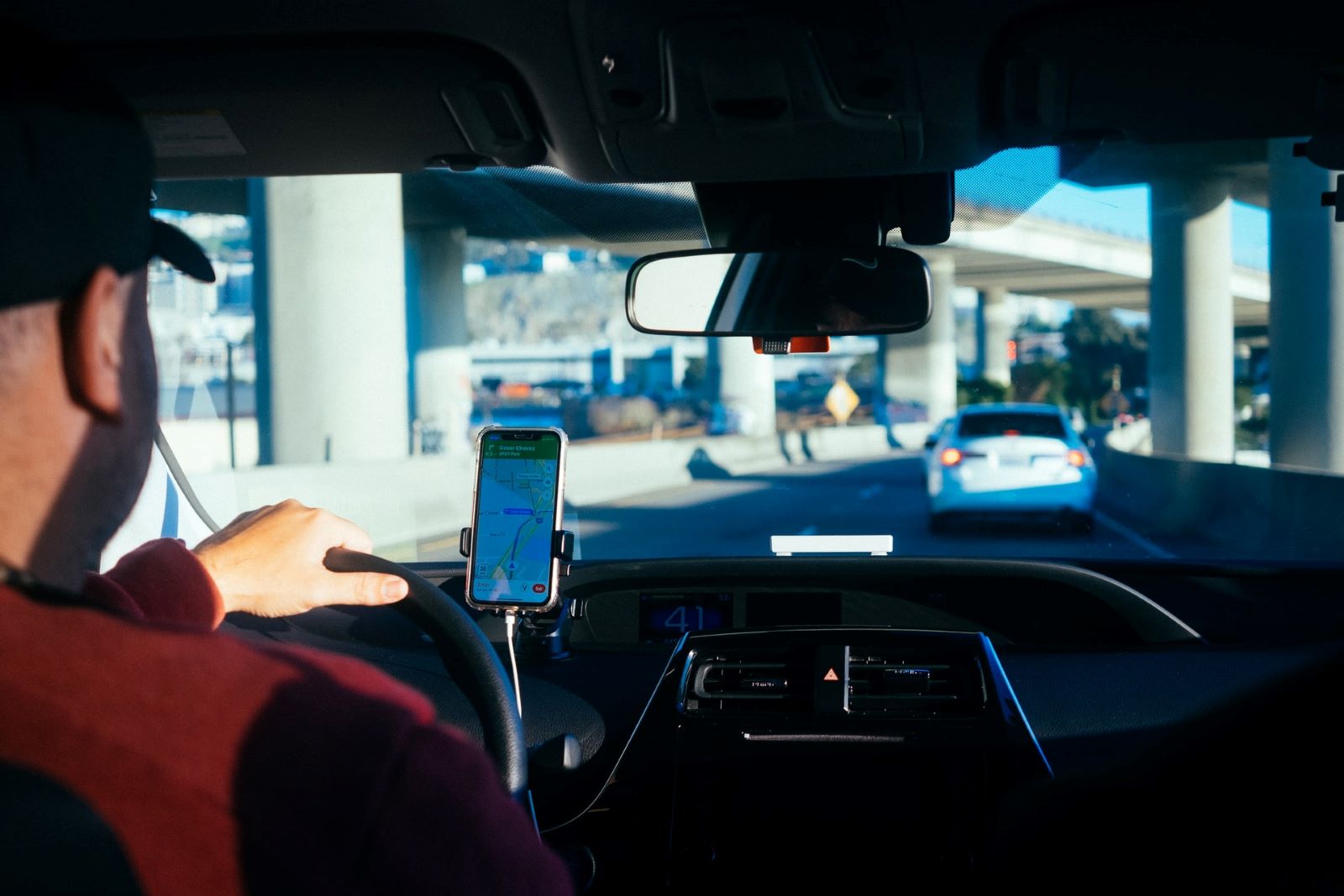 View from the backseat of a man in a hat driving a rideshare car. His cell phone is mounted to the dashboard with directions, and he's driving in a city
