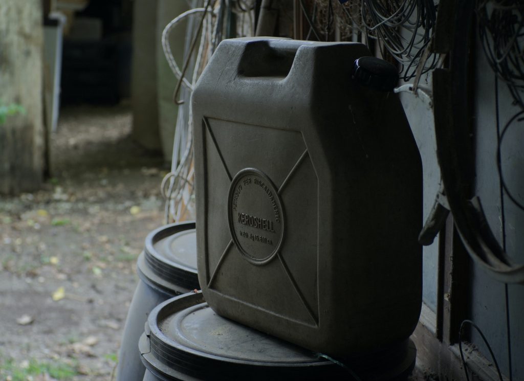 A plastic fuel can full of gasoline, sitting on a barrel in a shed.