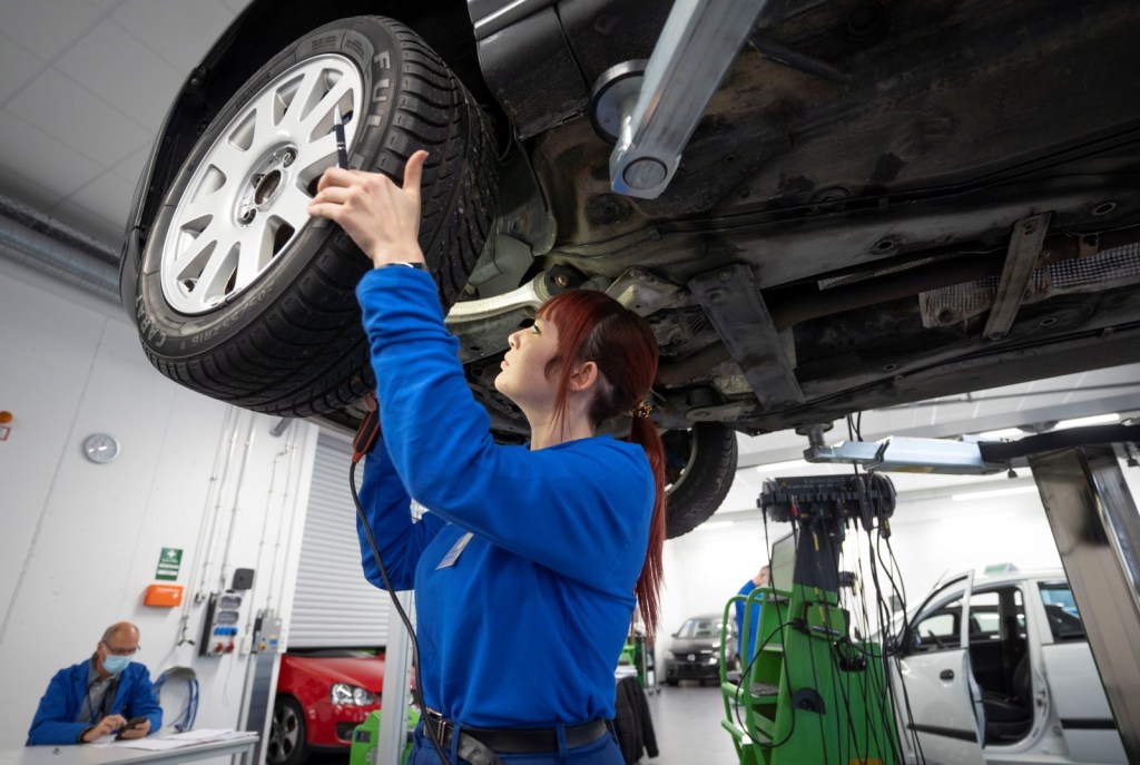 A female mechanic wearing a blue jumpsuit looks for brake problems on a car that's up on a lift