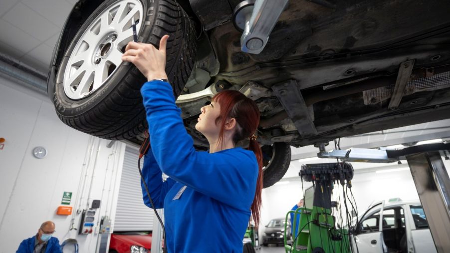 A female mechanic wearing a blue jumpsuit looks for brake problems on a car that's up on a lift