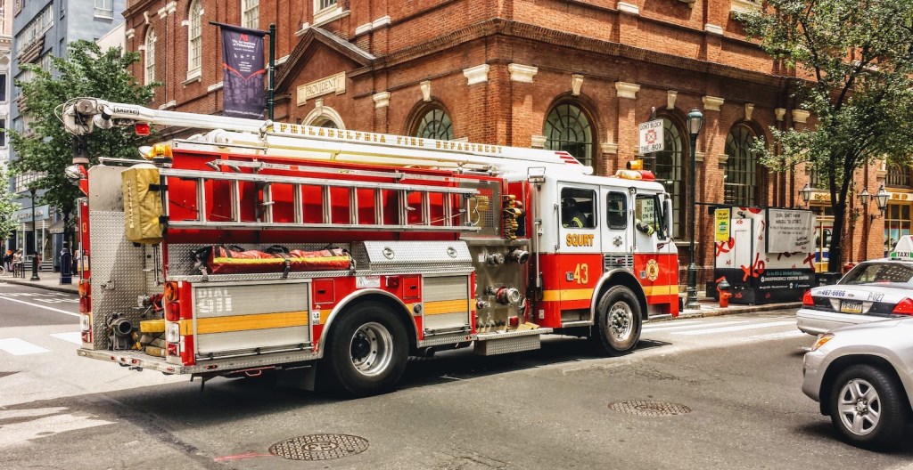 A used fire truck navigating an intersection in a crowded city, in front of an old brick building.