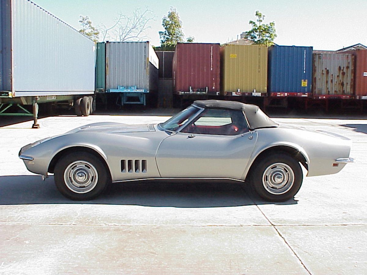 A silver 1968 Chevy Corvette C3 Stingray parked in front of box trailers in the background.