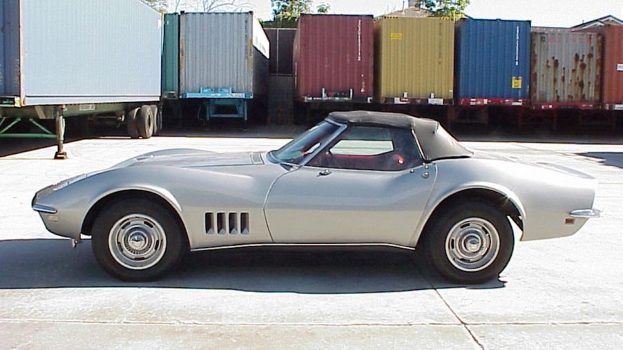A silver 1968 Chevy Corvette C3 Stingray parked in front of box trailers in the background.