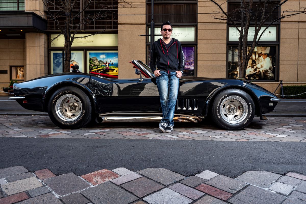 A Japanese business owner, wearing a black sweatshirt, jeans, and black sneakers, leans against his black 1969 Chevrolet Corvette
