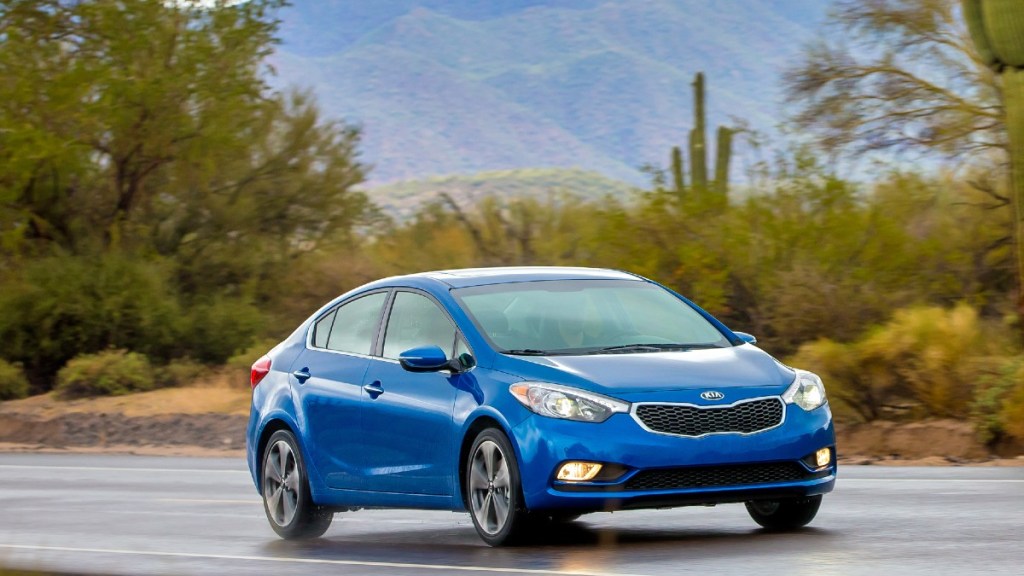 A blue 2016 Kia Forte drives along a desert road with mountains in the background 
