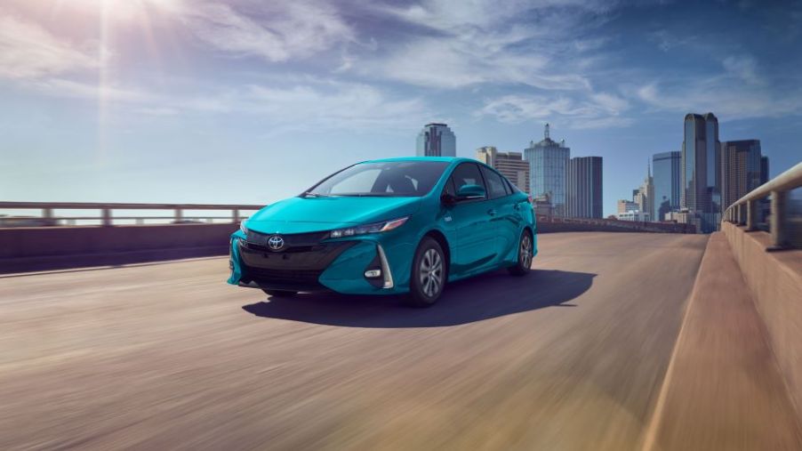 A blue 2022 Toyota Prius Prime driving down a road with a city in the background.