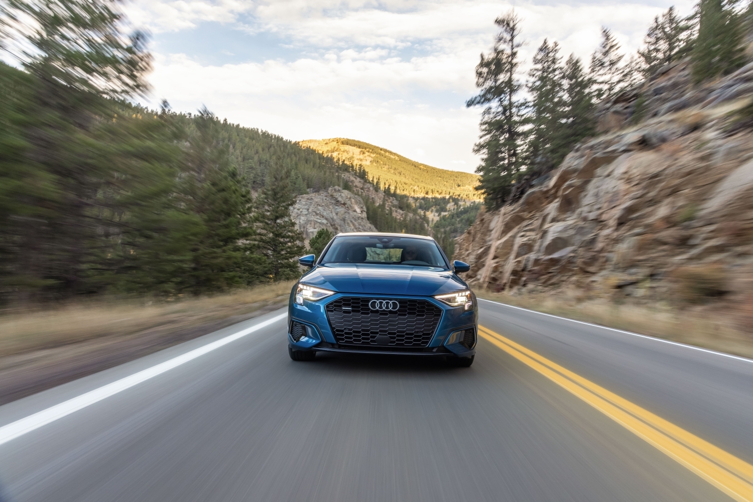 Front view of a blue 2022 Audi A3 sedan driving in the mountains; action shot, it's moving fast along a paved road