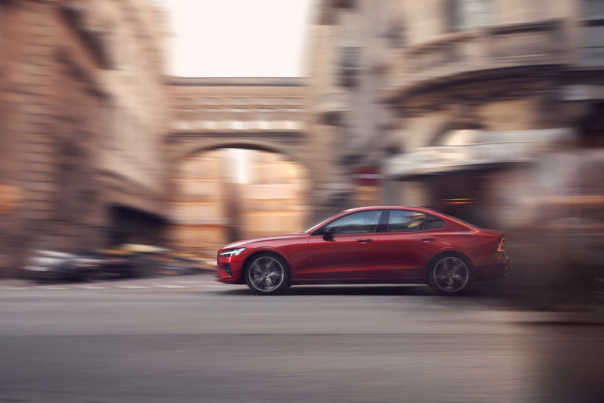 A red Volvo S60 plug-in hybrid, the best luxury PHEV, drives along a city street surrounded by brick buildings