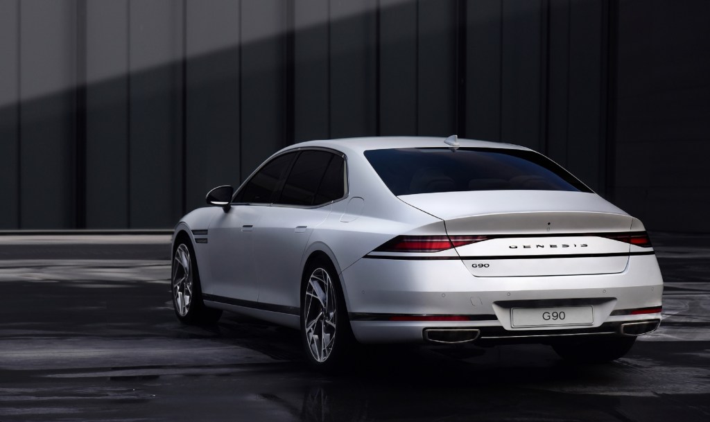 The rear 3/4 view of a white 2023 Genesis G90 by a gray concrete wall