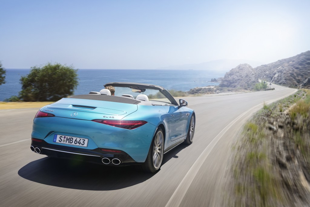 The rear 3/4 view of a blue 2023 Mercedes-AMG SL 43 driving down a mountain next to a body of water