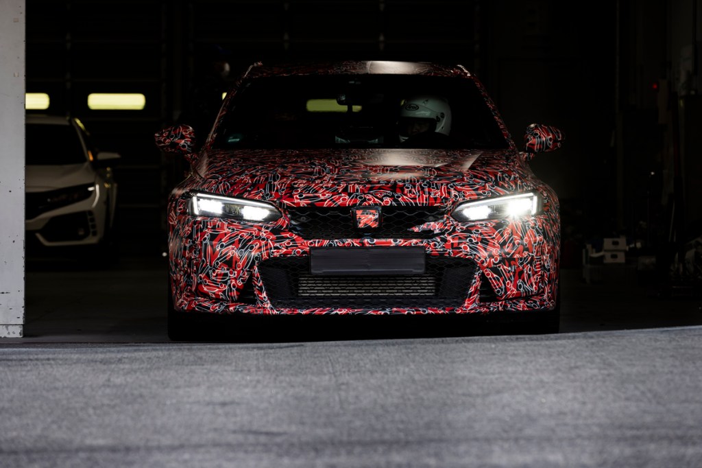 A heavily camouflaged 2023 Honda Civic Type R rolls out from a garage with a racecar driver in the driver's seat
