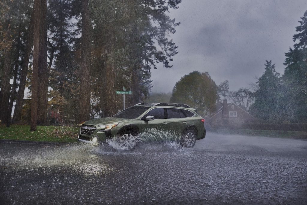A green updated 2023 Subaru Outback splashing through a puddle as it makes a sharp turn in the rain.