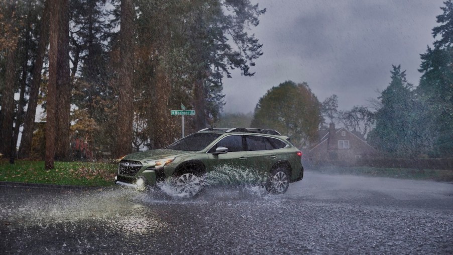 A green updated 2023 Subaru Outback splashing through a puddle as it makes a sharp turn in the rain.