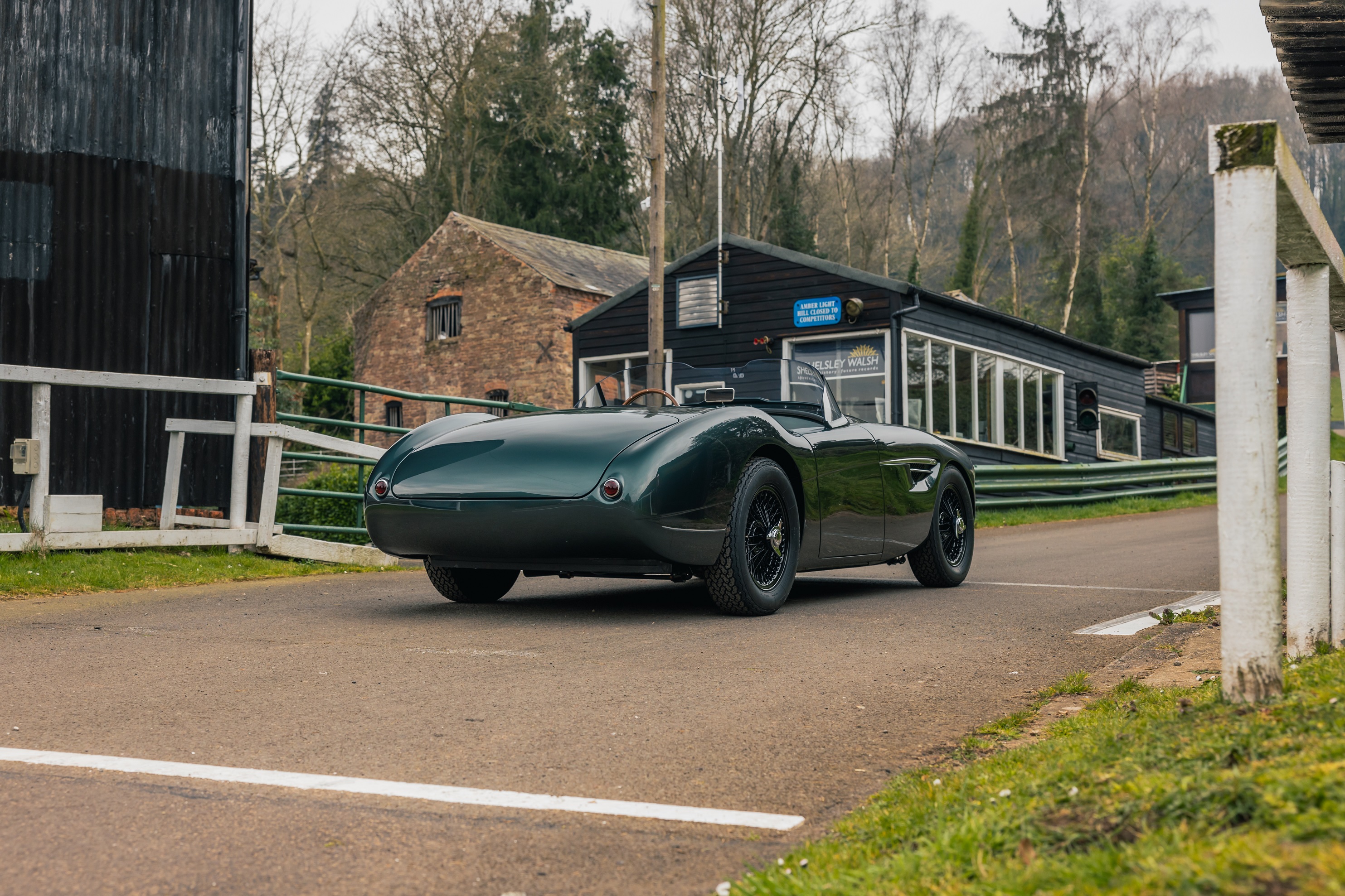 The rear 3/4 view of a dark-green Caton Austin-Healey 100 restomod on a British countryside track