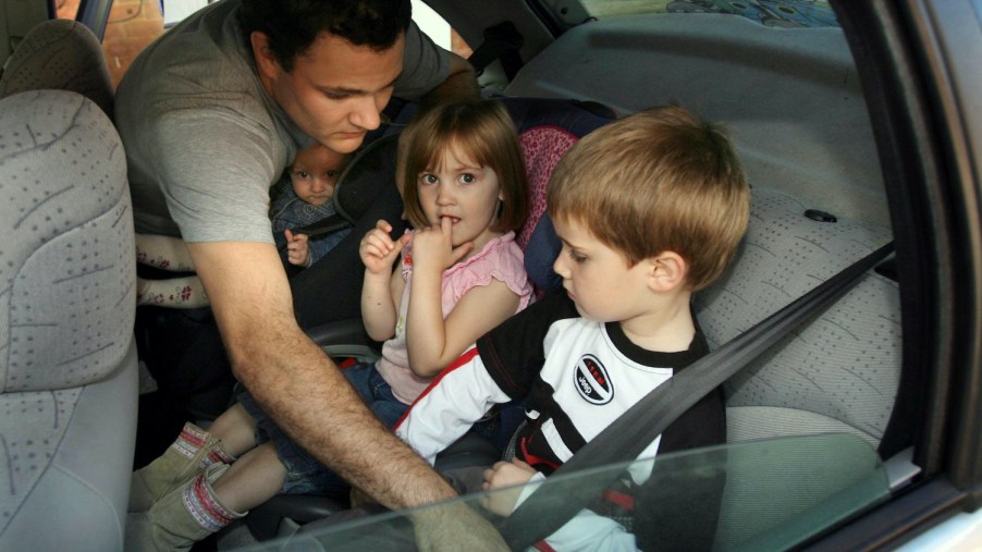 A dad buckles his kids into the back seat of a car.