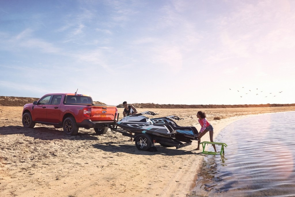 A red compact truck, the Ford Maverick drops off some jet skis at a beachfront.