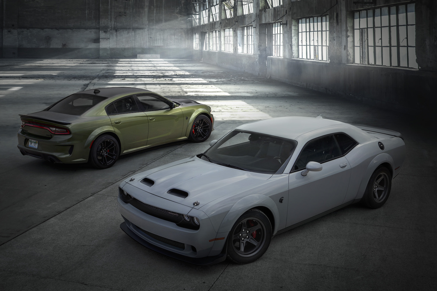 A green Dodge Charger and white Dodge Challenger parked in a warehouse for a promo photo. 