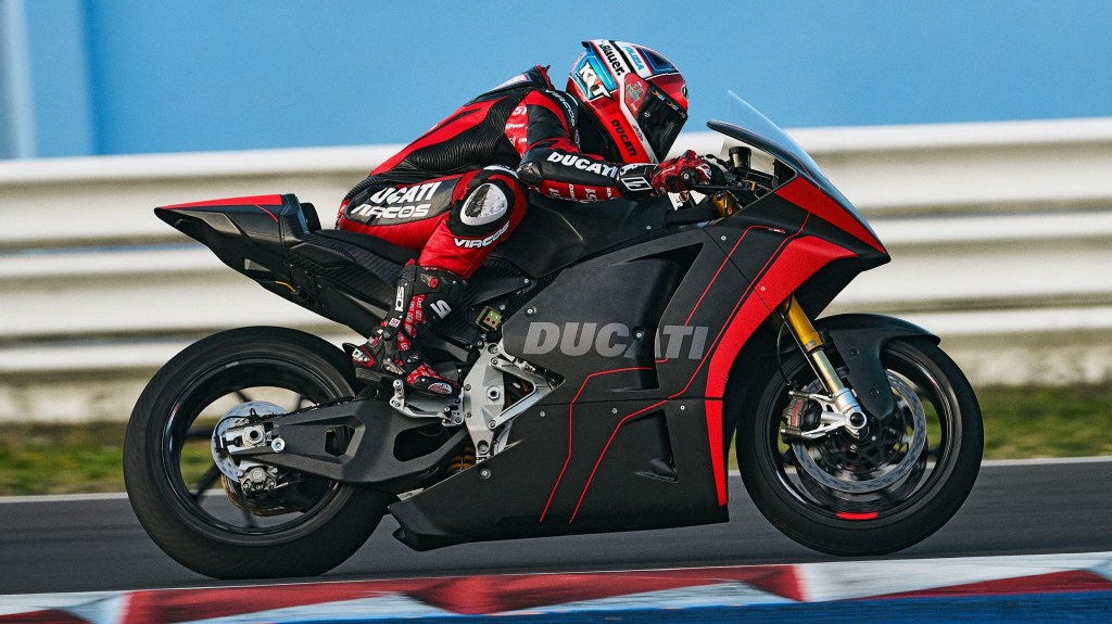 The side view of a black-and-red-clad test rider taking the red-and-black Ducati V21L MotoE electric motorcycle prototype around a racetrack