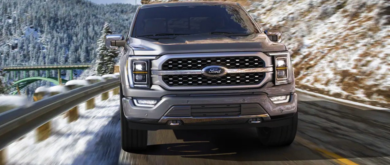 A gray 2022 Ford F-150 Platinum full-size pickup truck is driving on the road with snowy mountains in the background.