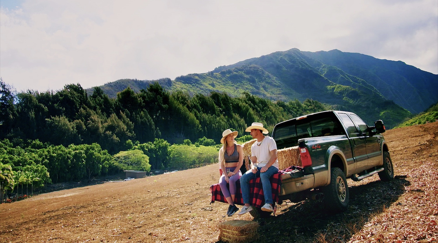 Couple sitting on the tailgate of a Ford F-150 pickup truck, a range of mountains in the background.