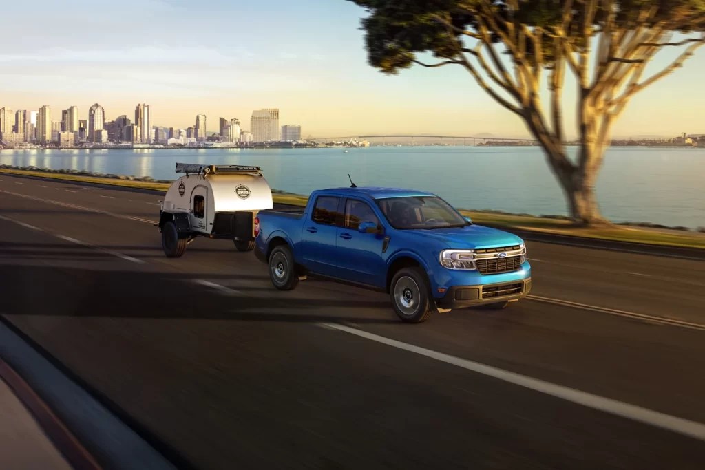 A blue Ford Maverick tows a small trailer, demonstrating its capability as a small truck.  He is driving along a riverside road with an urban background.