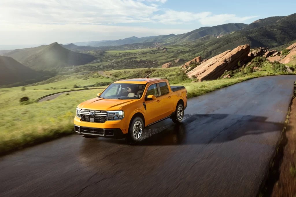 A yellow Ford Maverick drives on a scenic road as a 2022 small pickup truck.