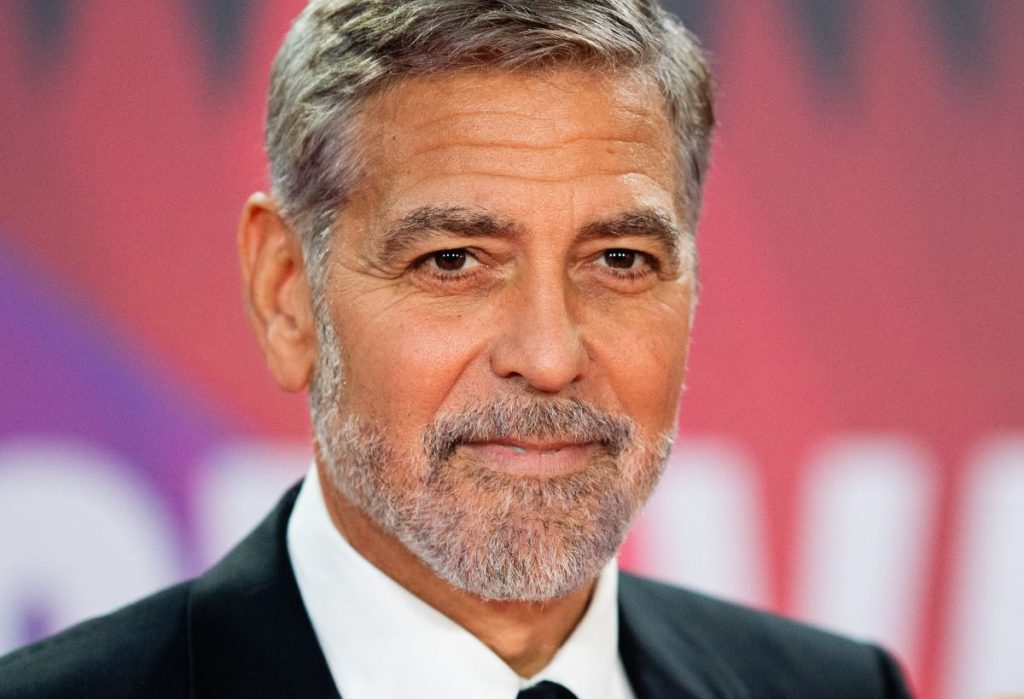 George Clooney standing against a blurred pink and white background. 