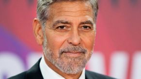 George Clooney standing in front of a pink and white background that is blurred.