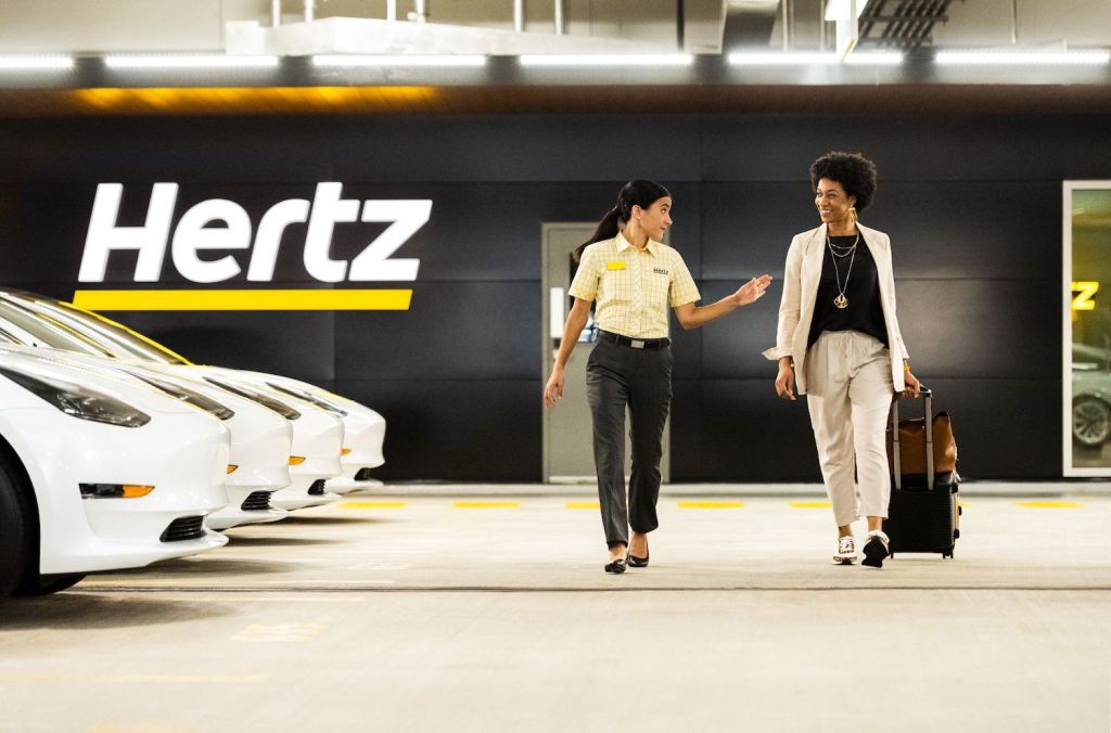Two women walking by a row of Hertz rental Tesla Model 3 cars in a commercial.