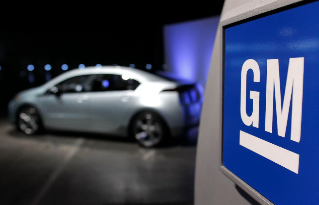 GM corporate sign with a Chevy Volt plug-in hybrid parked in the background.