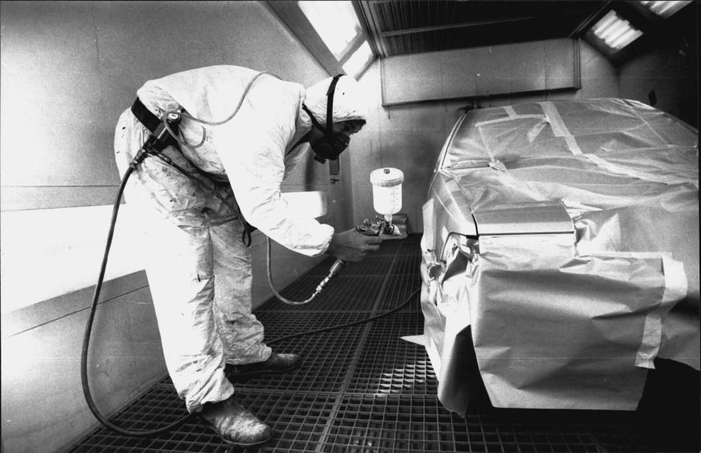 A technician in a white suit bending over the spray paint on the side of a car.