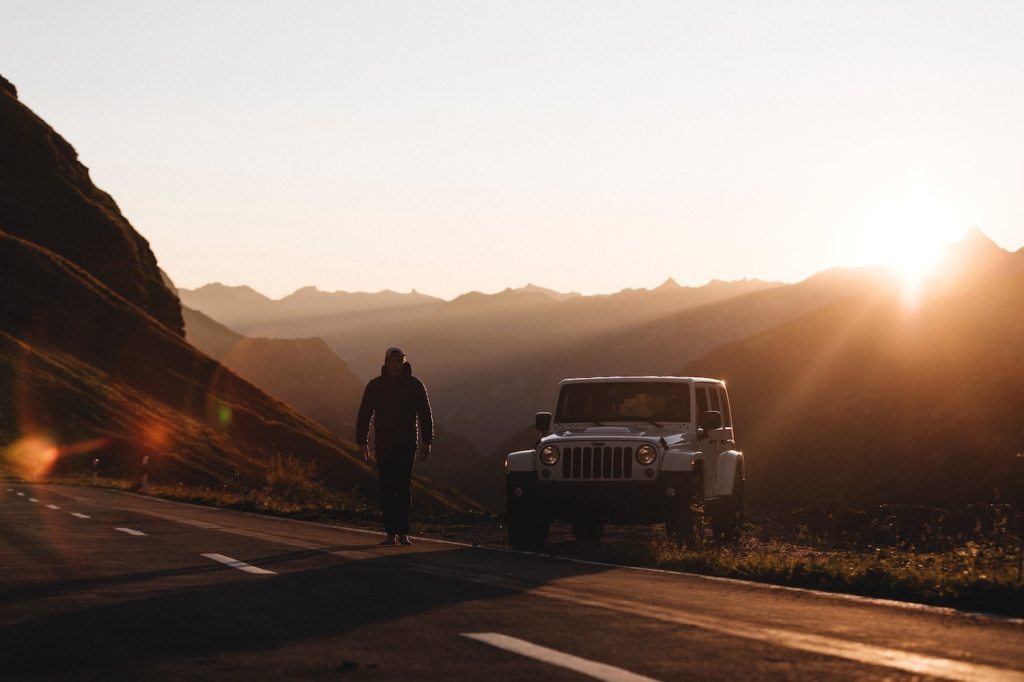 Man standing next to a Jeep Wrangler on a deserted road with the sunset above a row of mountains in the background.