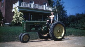 A kid drive a John Deere tractor in front of his brick house in 1946.