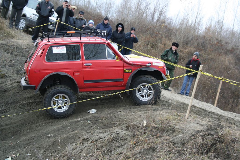 A Russian Lada Niva with a snorkel navigates a dirt trail as people watch in the background.