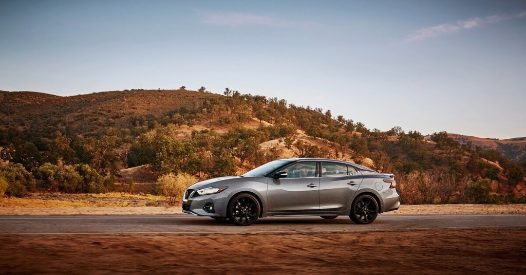 A gray Nissan Maxima parks next to an autumn scene. 