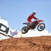 A person performing a jump on a dirt bike in protective gear on a dirt track outdoors.