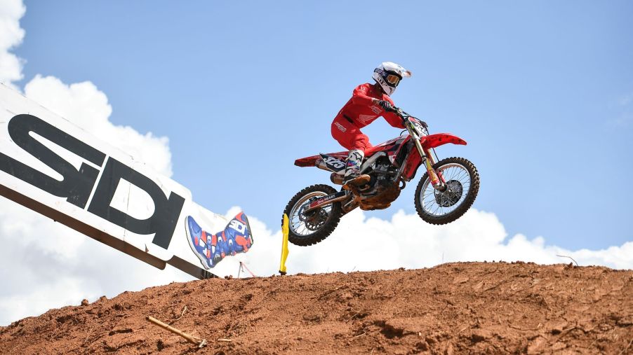 A person performing a jump on a dirt bike in protective gear on a dirt track outdoors.