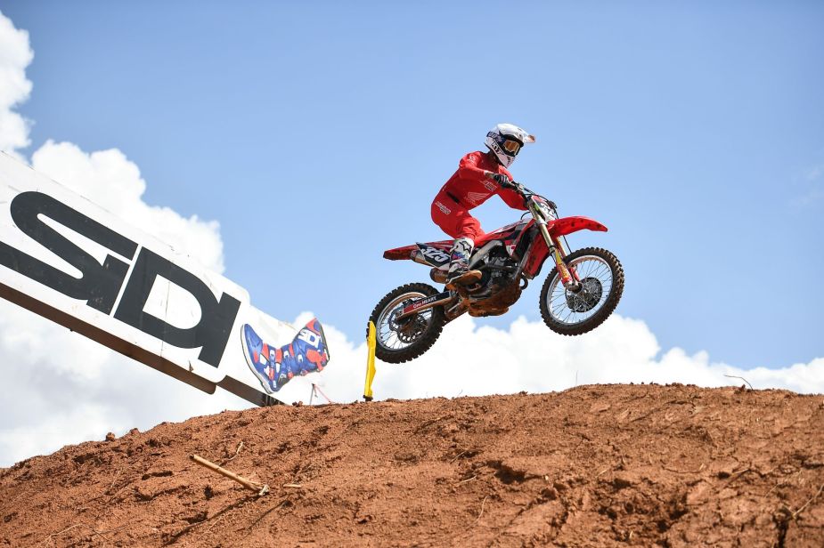 A person performing a jump on a dirt bike in protective gear on a dirt track outdoors.