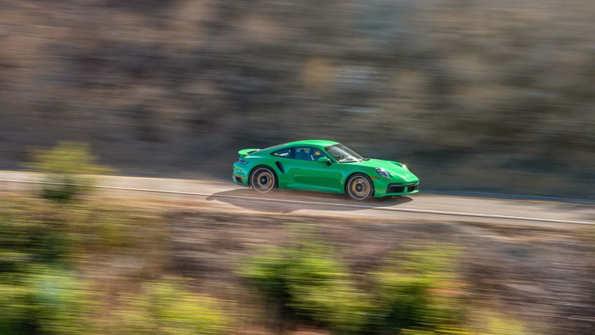 A 2022 Python Green Porsche 911 Turbo S speeds along a cliffside road during bright sunlight