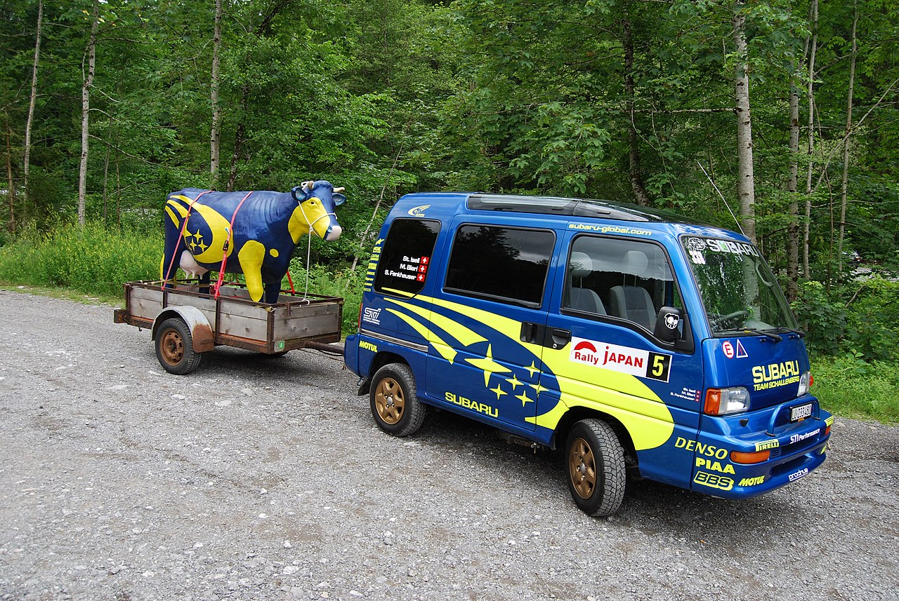 A Subaru Sambar wears Subaru rally paint and hauls a small trailer behind it as a Kei car.