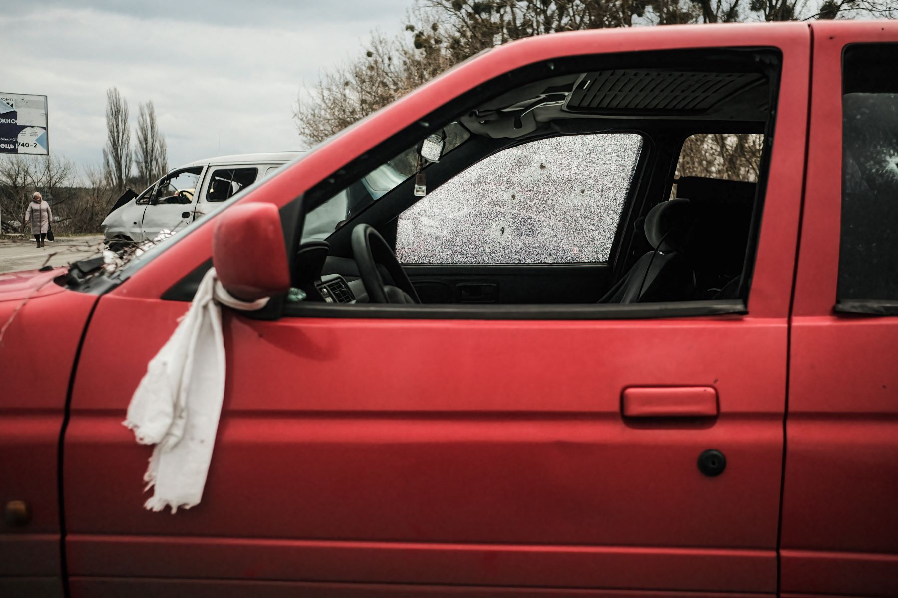 An abandoned car riddled with bullet holes with a white towel tied around the driver's side mirror