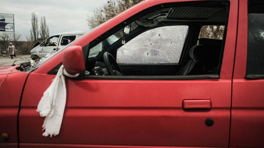 An abandoned car riddled with bullet holes with a white towel tied around the driver's side mirror