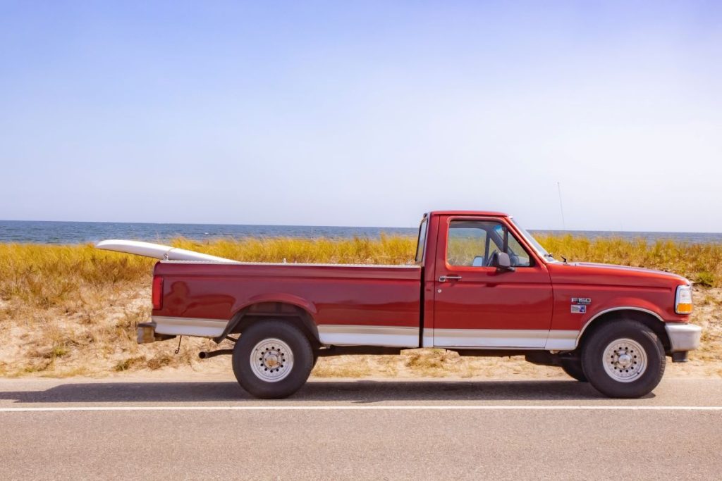 A well-kept classic red Ford F-150 small pickup truck, parked in front of the beach with a surfboard strapped to the bed. 