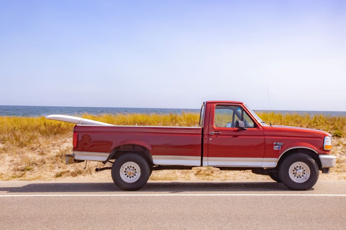 Well-maintained, classic Ford F-150 small pickup truck in red, parked in front of a beach with a surfboard strapped into the bed