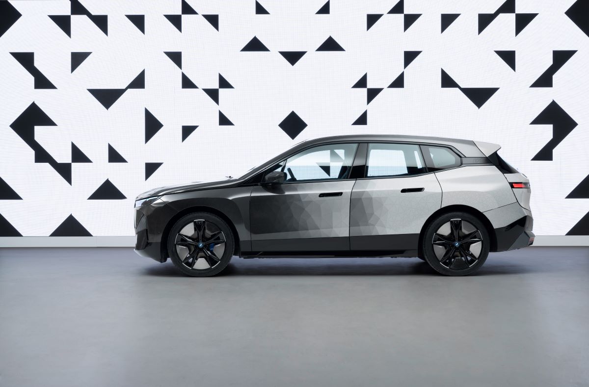 Side view of the BMW iX M60 Electric SUV with color-changing paint, with colors slowly changing from black to white front to back. The background is a black-and-white abstract pattern