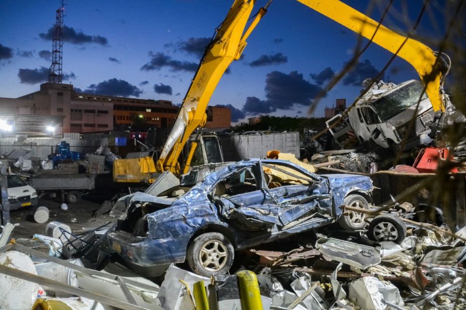 A yellow excavator pulls apart a busted old blue car at a junkyard