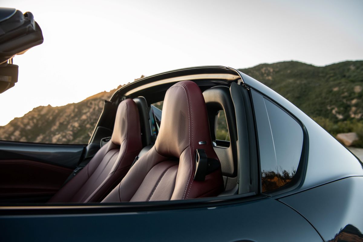 Close-up shot of the driver and passenger seats in a Mazda Miata with the top down. There's a small panel behind the seats to allow for air flow