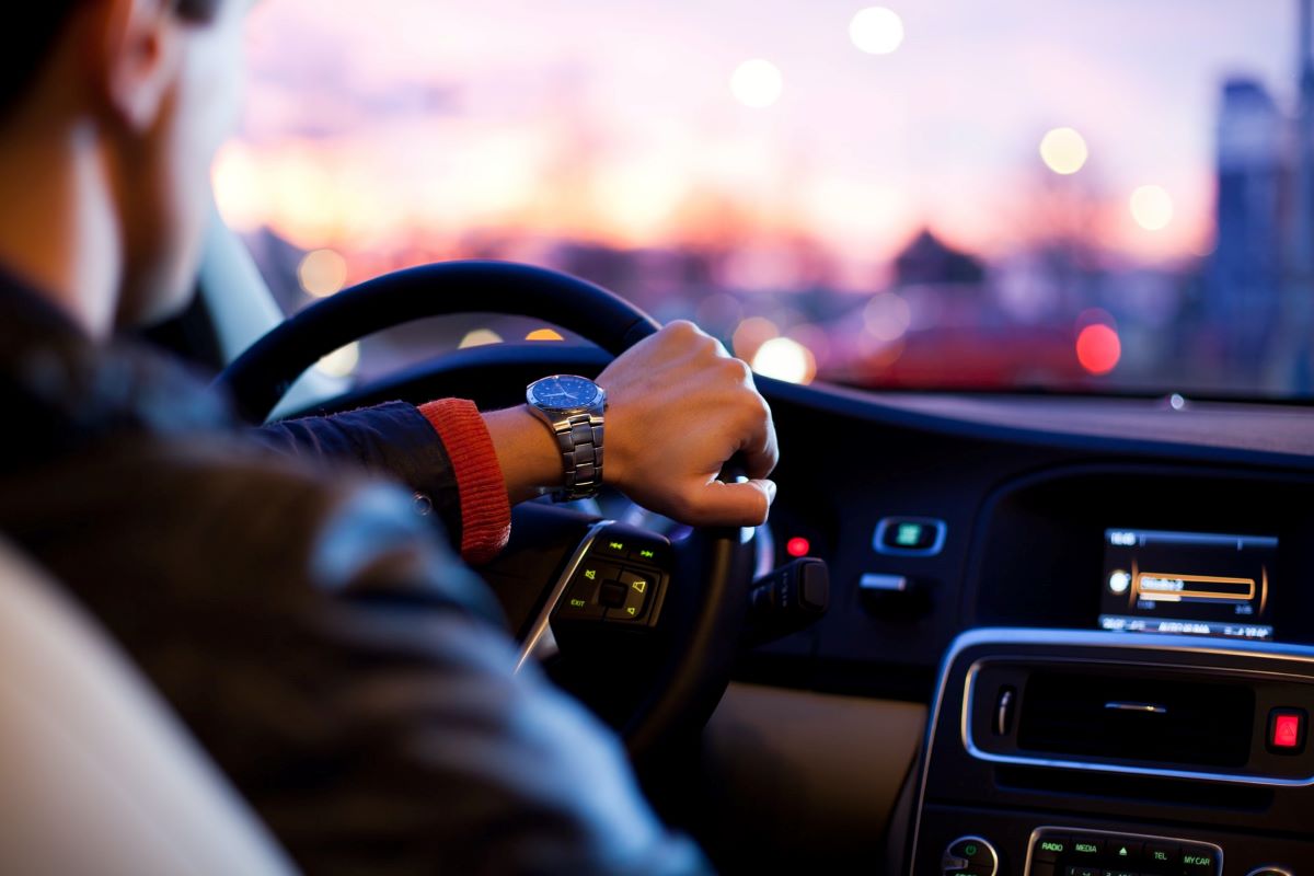Image of a man driving a car. The focus is on his left hand, which holds the wheel. He's wearing a watch, and there's music on the car radio
