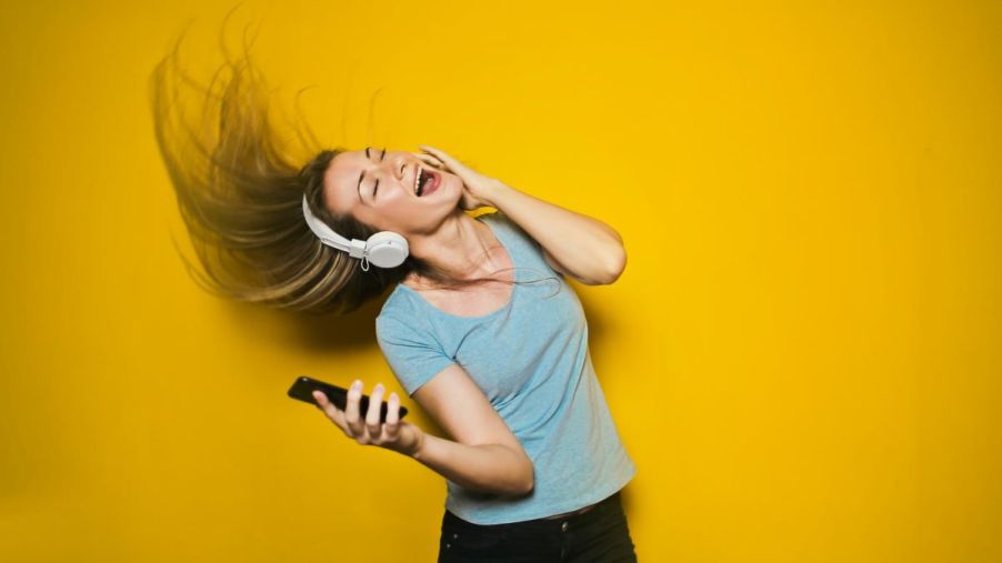 Blonde woman listening to upbeat music, dancing and whipping her hair. She's wearing white headphones, a green shirt, and black pants. She holds a smartphone in her right hand.