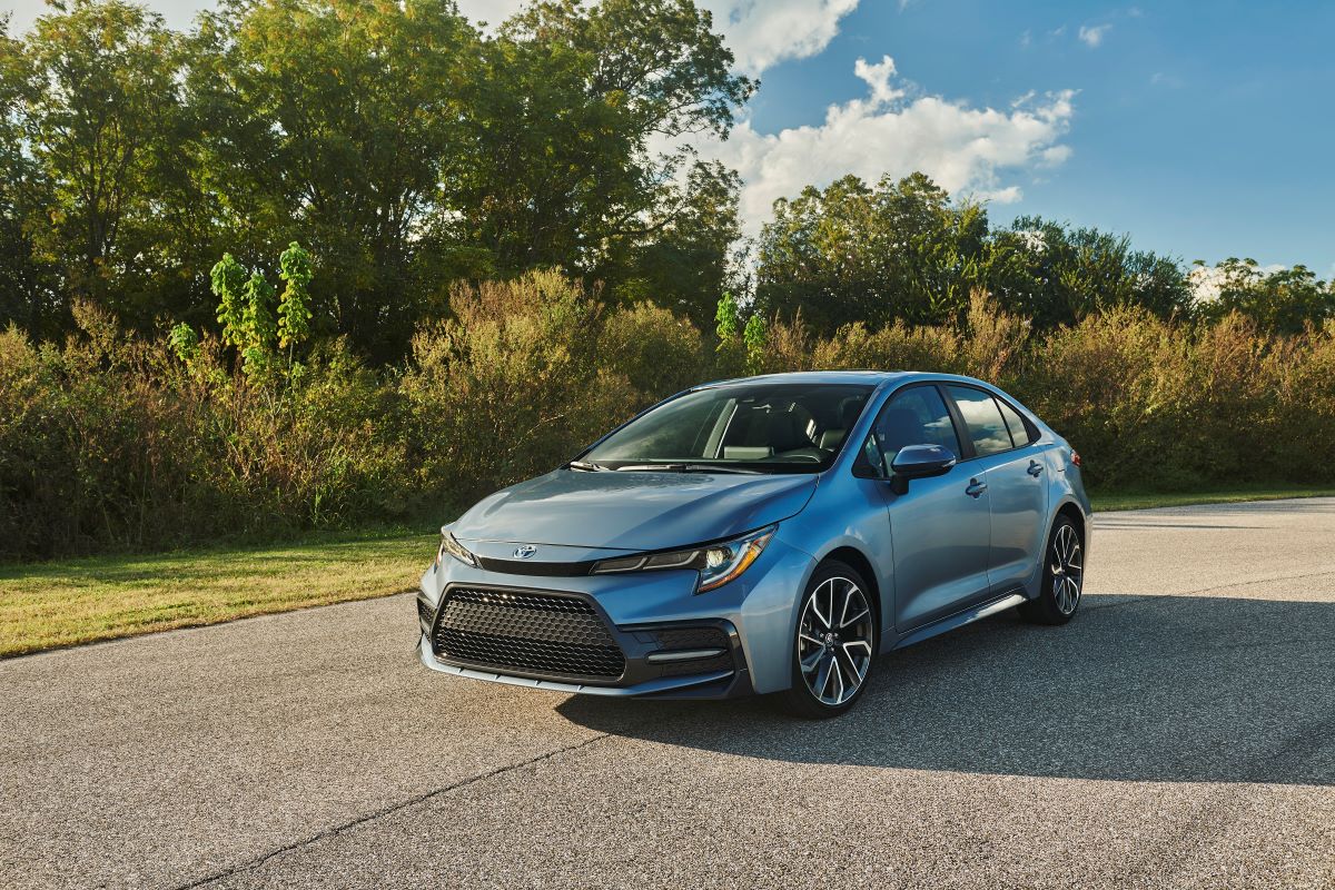 Used 2020 Toyota Corolla sedan in grey, parked on pavement in front of trees and bushes on a summer day